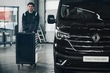 A technician in workwear moves a tool trolley next to a black Renault Trucks Trafic Red EDITION in a dealership workshop. This image highlights the technical expertise and professional maintenance services provided by the Renault Trucks network.